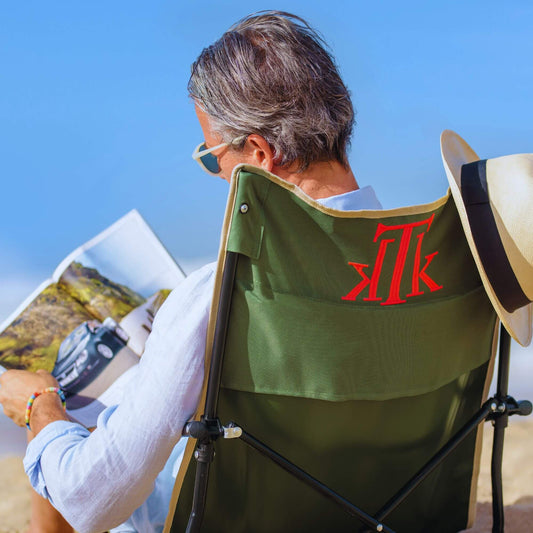 Hombre leyendo una revista sentado en una silla de playa plegable verde KTK Kontiki sobre la arena, con sombrero de verano colgado, disfrutando del mar con estilo y comodidad.