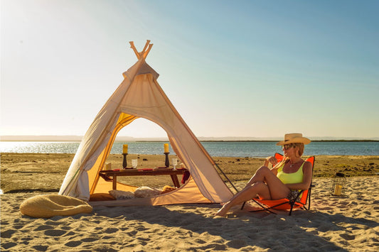 Mujer sentada en silla de playa naranja plegable KTK KONTIKI junto a una tienda tipi frente al mar al atardecer, disfrutando del verano con estilo y comodidad.