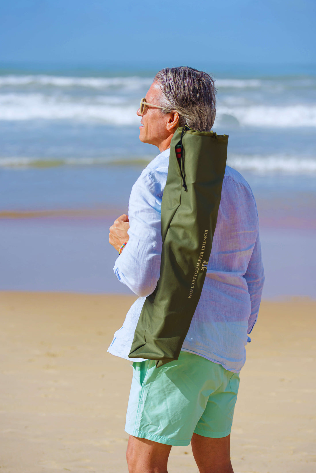 Hombre en la playa con silla de playa plegable KTK Kontiki en bolsa verde al hombro, destacando su portabilidad, diseño ligero y estilo elegante para verano 2025