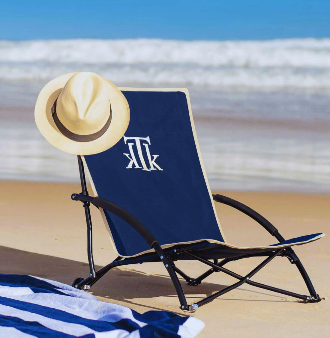 Silla de playa plegable premium KTK Kontiki en azul marino con estructura ligera y diseño ergonómico, ideal para vacaciones de lujo en la costa. Accesorio playero chic con sombrero elegante sobre la arena frente al mar Mediterráneo en España.