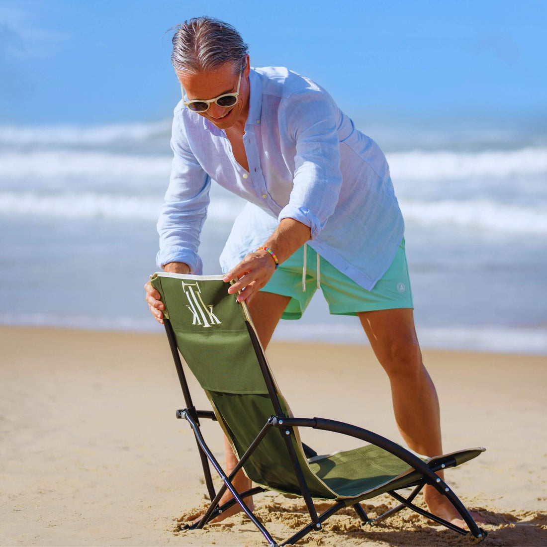 Hombre elegante montando una silla de playa plegable verde KTK Kontiki en la orilla del mar, disfrutando de unas vacaciones con estilo y confort en la playa, silla portátil ligera y ergonómica ideal para escapadas premium de verano 2025.