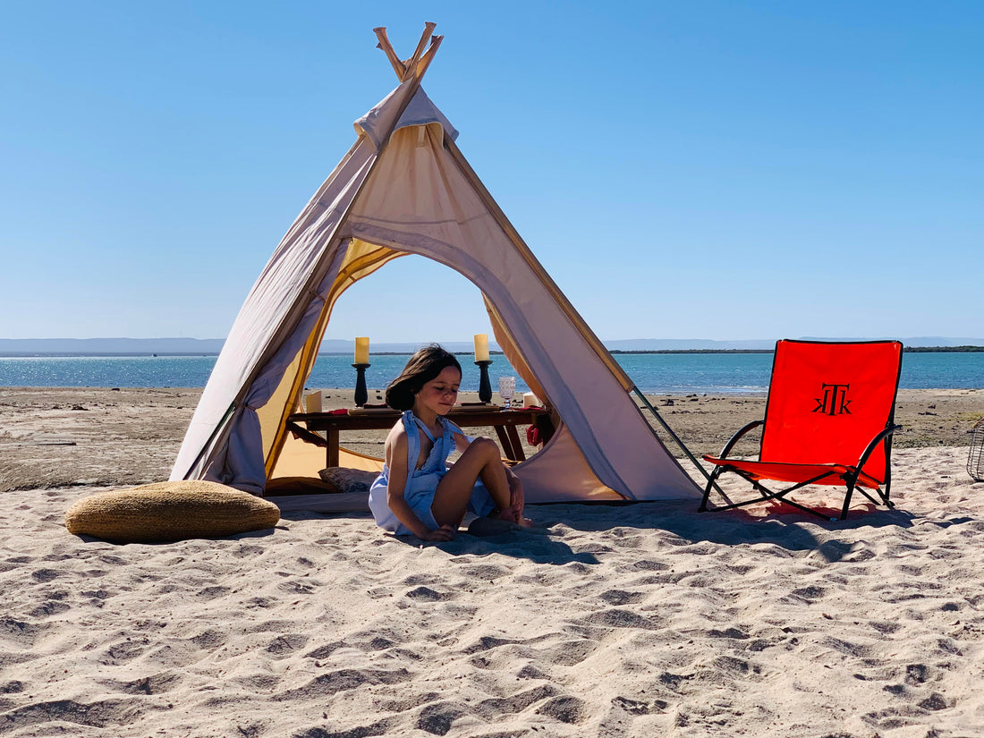 Niña jugando en la playa junto a silla de playa plegable portátil KTK Kontiki color naranja, diseño ergonómico ideal para familias, escena veraniega con decoración boho y vista al mar.